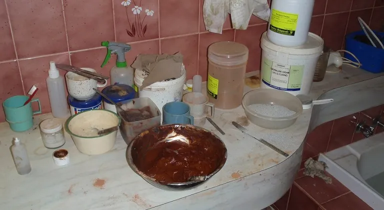 Cluttered work surface in a manufacturing or preparation area, with multiple open containers, bowls, utensils, bottles and visible residue spread across the bench.